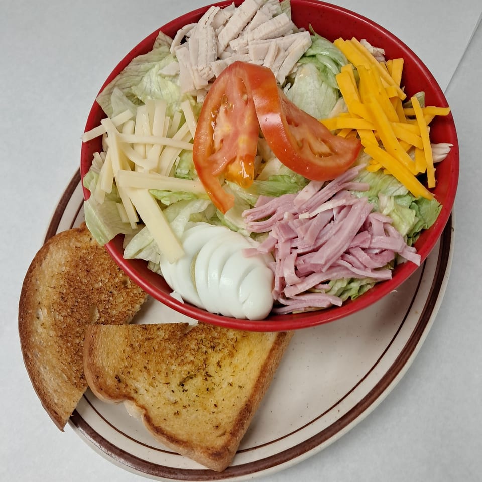 Chef salad with turkey, ham, cheese, tomato, and egg in a red bowl, with toast on a plate.